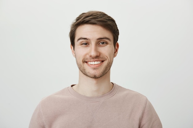 Free photo confident attractive caucasian guy in beige pullon smiling broadly while standing against gray