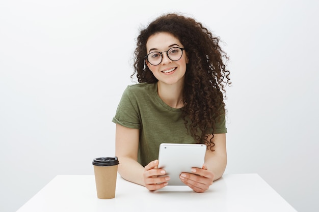 Confident attractive curly-haired brunette in trendy eyewear, sitting at table, drinking tea or coffee and holding digital tablet