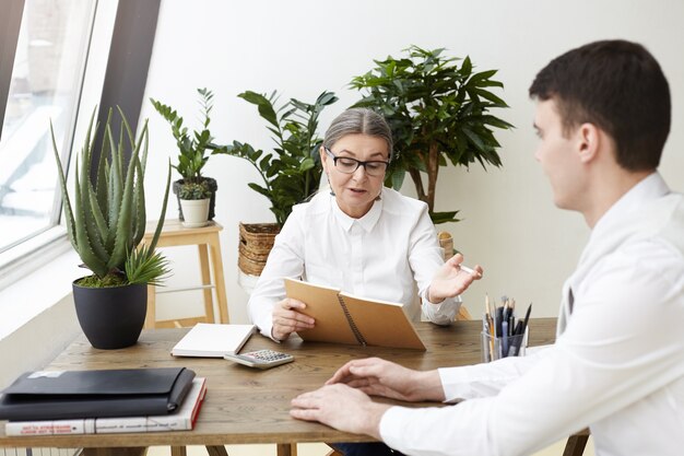 Confident attractive woman CEO in her fifties holding copybook while posing questions about job qualification, experience and skills during interview with brunette male applicant. Film effect