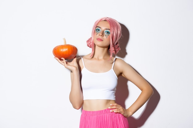 Уверенная в себе привлекательная женщина в розовом парике с ярким макияжем держит тыкву на праздновании Хэллоуина ...