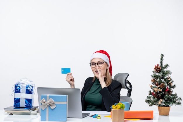 Free photo confident attractive woman with santa claus hat and wearing eyeglasses sitting at a table and holding bank card and gossiping in the office