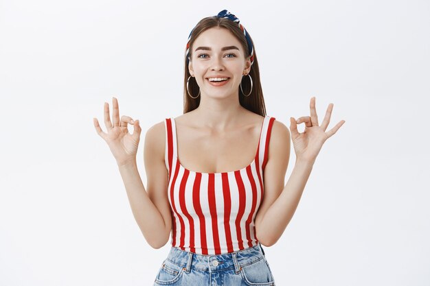 Confident attractive young woman in striped top and headband showing okay gesture with both hands and smiling