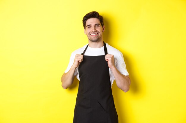 Confident barista in black apron standing against yellow background. Waiter smiling and looking happy