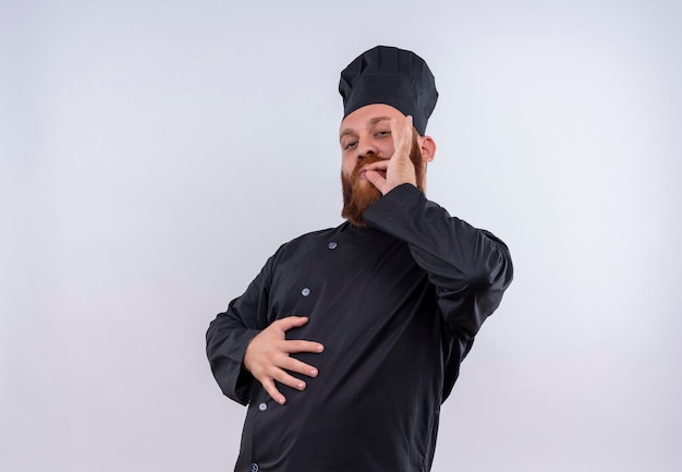 A confident bearded chef man in black uniform holding hand on his stomach and showing ok gesture on a white wall