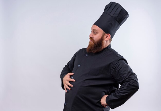 A confident bearded chef man in black uniform keeping hand on his stomach while holding hand on his waist on a white wall