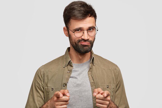 Confident bearded man points with both index fingers, selects something, has dark stubble, stands alone against white wall. Attractive self assured ushaven male expresses his choice