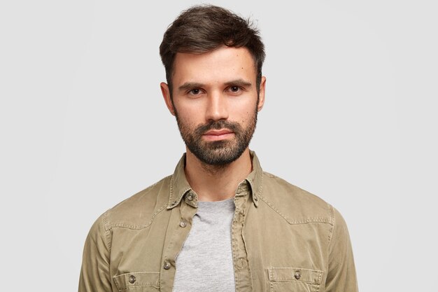 confident bearded man with dark hair, has serious facial expression, thinks about future job, dressed in fashionable shirt