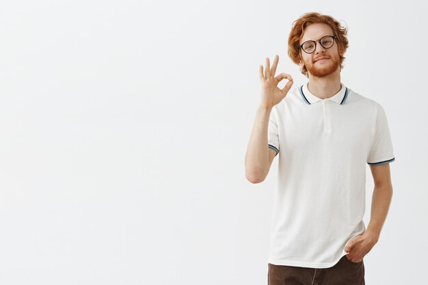 Confident bearded redhead guy posing against the white wall with glasses