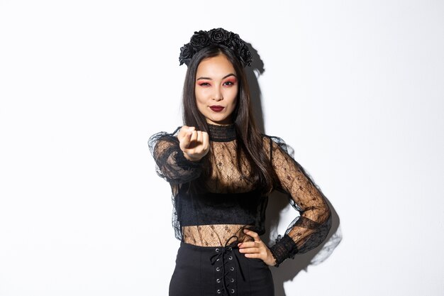 セクシーな魔女の衣装を着た自信のある美しいアジアの女性があなたを誘惑し、白い背景の上に立って、ハロウィーンを祝い、ゴシックドレスを着て、伸ばした指で近づいてください。
