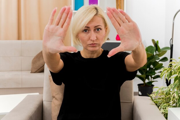 Confident beautiful blonde russian woman sits on armchair stretching hands out looking at camera inside the living room