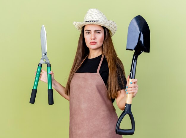 Уверенная красивая девушка-садовник в униформе в садовой шляпе, держащая ножницы с лопатой на оливково-зеленом фоне