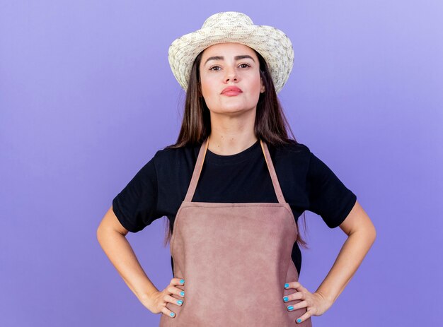 Confident beautiful gardener girl in uniform wearing gardening hat putting hands on hip isolated on blue