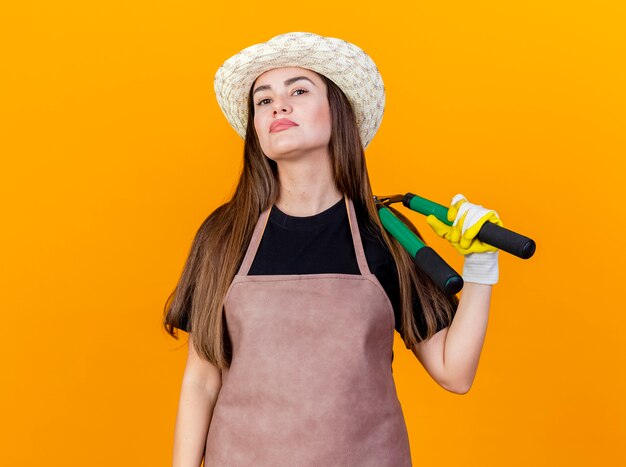 オレンジ色の背景で隔離の肩にバリカンを保持している手袋と制服と園芸帽子を身に着けている自信を持って美しい庭師の女の子