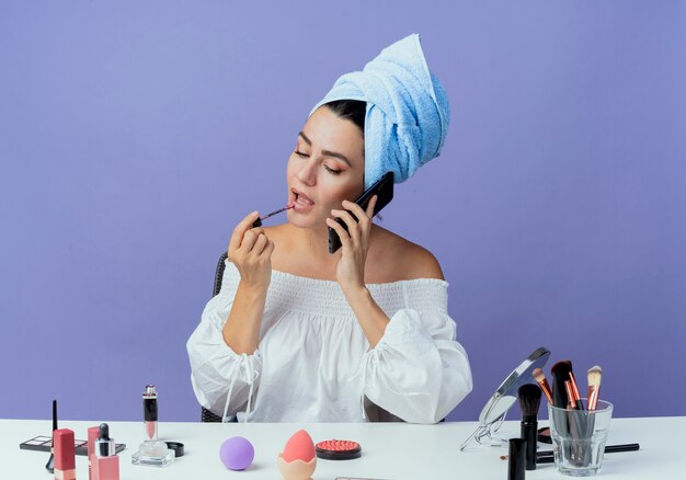 Confident beautiful girl wrapped hair towel sits at table with makeup tools holding and applying lip gloss talking on phone isolated on purple wall