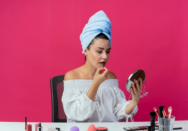 Confident beautiful girl wrapped hair towel sits at table with makeup tools holding and applying lipstick looking at mirror isolated on pink wall