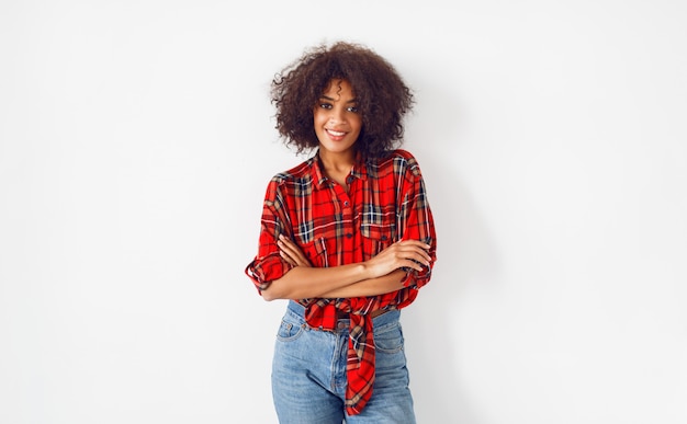 Ragazza nera sicura che posa sopra il fondo bianco. indossa una camicia a scacchi rossa. blue jeans.