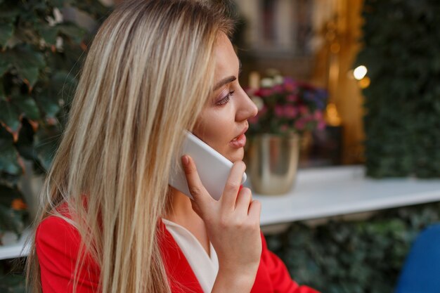 Уверенная белокурая деловая женщина в красном стильном пиджаке разговаривает по телефону mobyle и сидит в городском кафе
