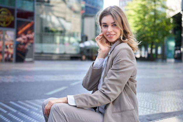 Confident blond businesswoman in suit smiles at camera sits outdoors near office buildings