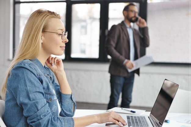 Confident blonde female secretary uses laptop while boss has phone conversation