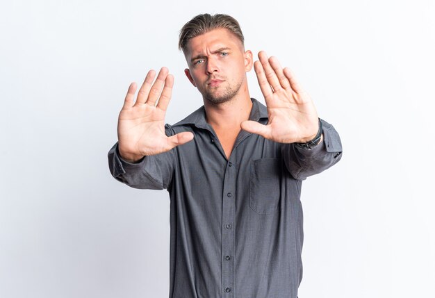 Confident blonde handsome man gesturing stop sign isolated on white wall with copy space