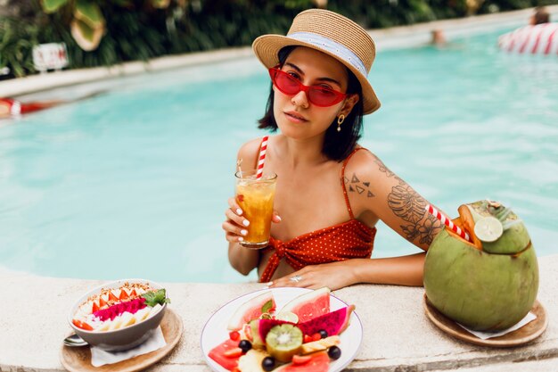 Confident  brunette tan woman in red cat eyes sunglasses   relaxing in   pool  with plate of exotic fruits  during tropical vacation.  Stylish tattoo.