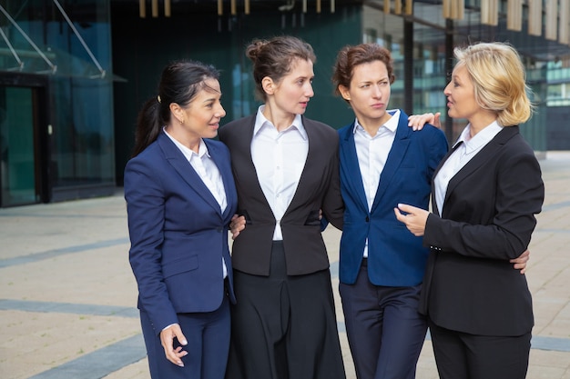 Confident business ladies standing together outdoors, hugging and talking. Businesswomen wearing suits meeting in city. Female team and teamwork concept