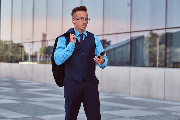 Fiducioso uomo d'affari vestito con un abito elegante tiene smartphone e giacca sulla spalla mentre si trova sullo sfondo del paesaggio urbano.