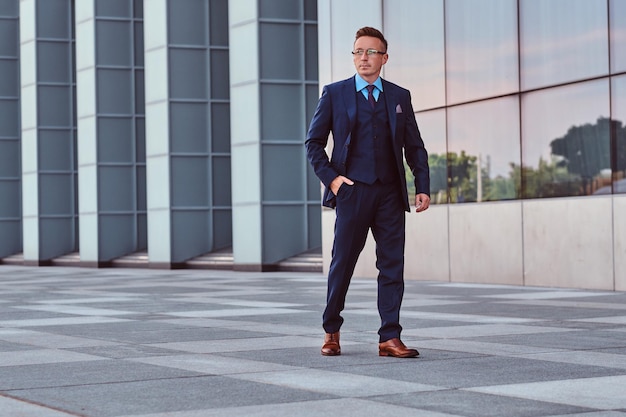 Free photo confident businessman dressed in an elegant suit looking away and holds hand in pocket while walk on the street outdoors against cityscape background.