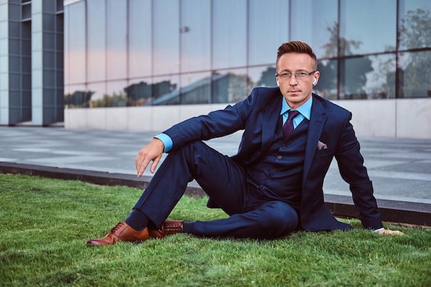 Confident businessman dressed in an elegant suit sitting on a green lawn against cityscape background.