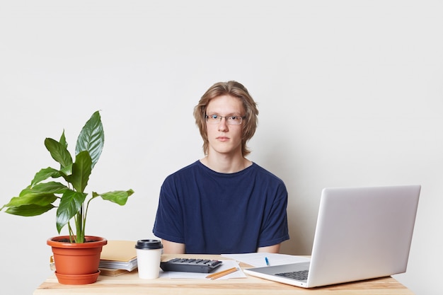 Confident businessman prepares annual report, calcualtes figures, uses modern laptop computer and calculator, drinks takeaway coffee, looks seriously into camera, isolated over white wall