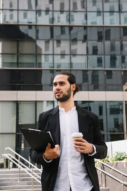 Free photo confident businessman standing in front of building holding clipboard and coffee cup