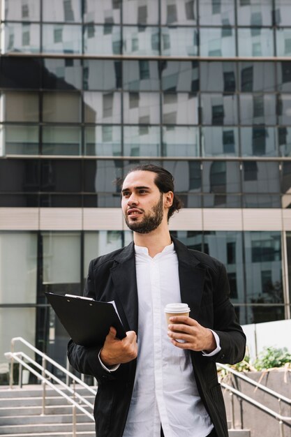 Confident businessman standing in front of building holding clipboard and coffee cup