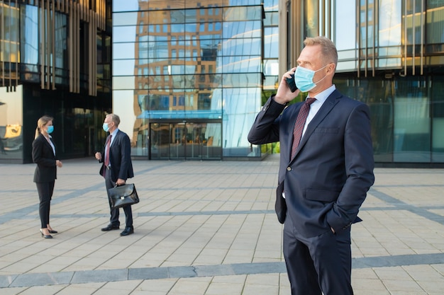 Confident businessman wearing mask and office suit talking on cellphone outdoors. Businesspeople and city building glass facade in background. Copy space. Business and epidemic concept