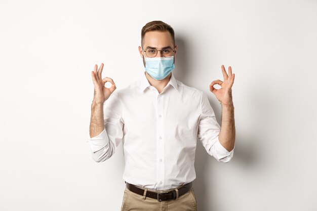   Confident businessman wearing medical mask and showing okay signs in approval, white background.