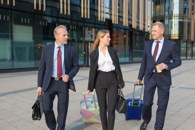 Confident businesspeople travelling with luggage, walking to hotel, wheeling suitcases, talking. Front view. Business trip or corporate communication concept