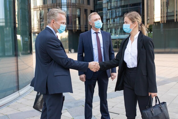 Confident businesswoman and middle-aged manager in facial masks handshaking outdoors. Successful employers greeting on street and working during coronavirus pandemic. Meeting and partnership concept