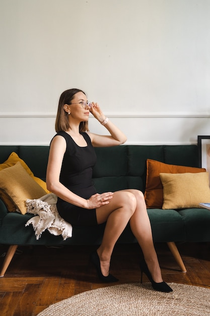 Уверенная деловая женщина в очках в черном классическом платье сидит в современной квартире на диване