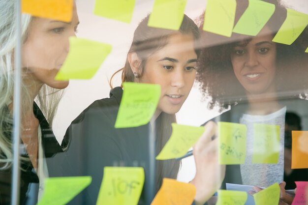 Confident businesswomen writing notes on stickers for startup. Successful experienced managers in suits meeting in conference room and planning strategy. Teamwork, business and management concept