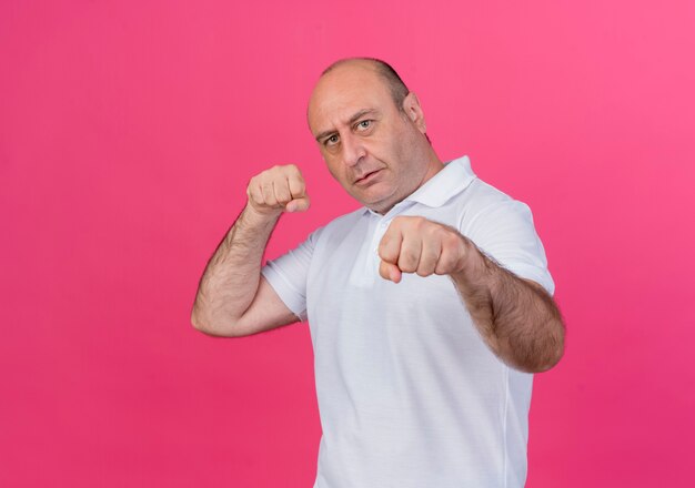 Confident casual mature businessman doing boxing gesture at camera isolated on pink background with copy space