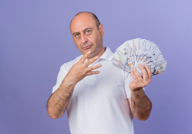 Confident casual mature businessman holding money and showing four with hand isolated on purple background with copy space