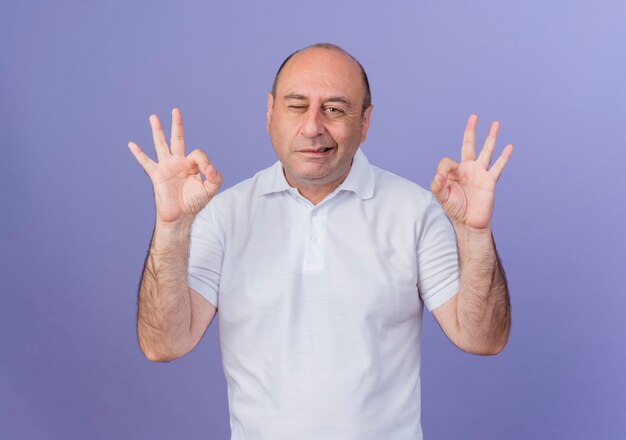 Confident casual mature businessman winking and doing ok signs isolated on purple background