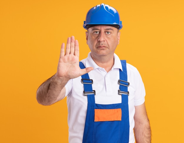 Бесплатное фото Уверенный кавказский взрослый человек-строитель в форме жестов стоп знак рукой на оранжевом