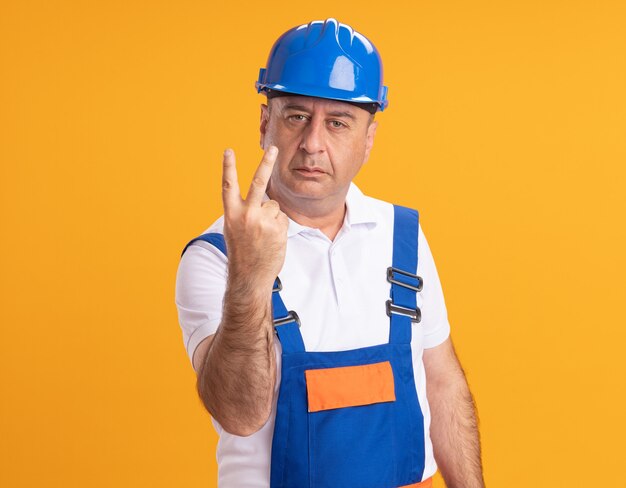 Confident caucasian adult builder man in uniform gestures two with finger on orange