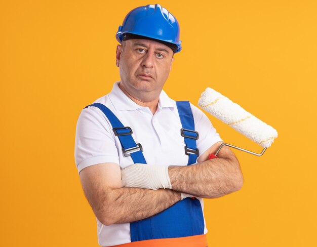 Уверенный кавказский взрослый человек-строитель в униформе стоит со скрещенными руками, держа роликовую щетку на оранжевом