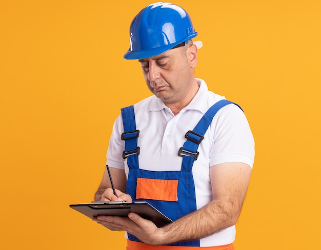 Confident caucasian adult builder man in uniform writes on clipboard with pencil on orange