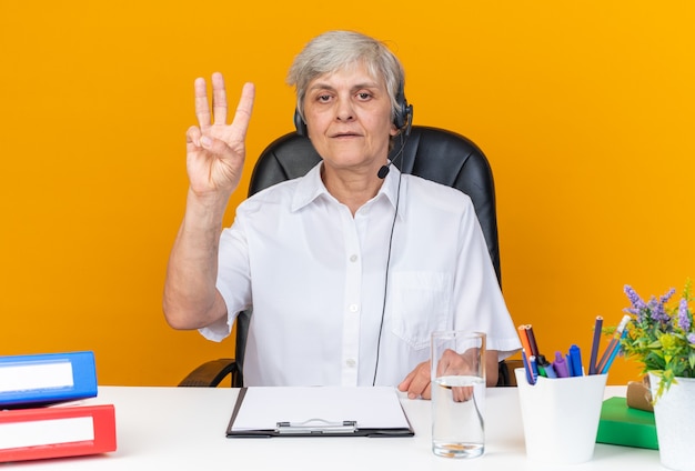 Confident caucasian female call center operator on headphones sitting at desk with office tools gesturing three with fingers 