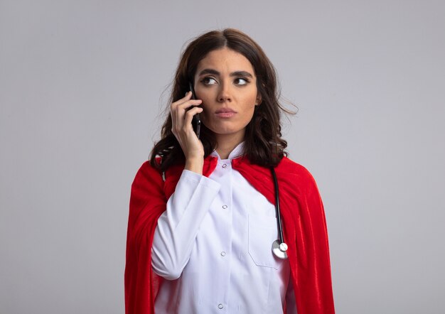 Confident caucasian superhero girl in doctor uniform with red cape and stethoscope talks on phone looking at side isolated on white wall with copy space