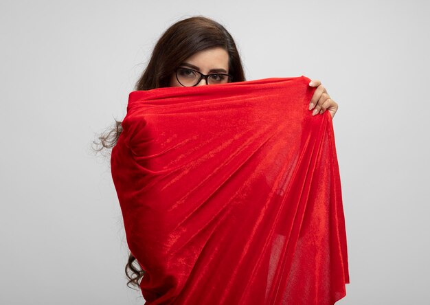 Confident caucasian superhero girl in optical glasses holds and looks at camera over red cape