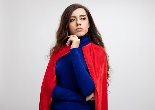 Уверенная кавказская девушка супергероя с красной накидкой держит руку и смотрит на сторону, изолированную на белой стене с копией пространства