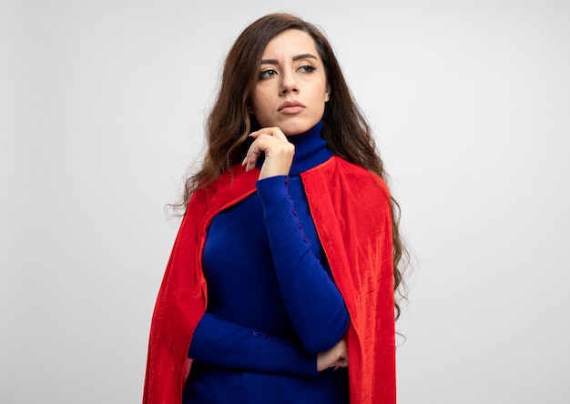 Confident caucasian superhero girl with red cape holds hand and looks at side isolated on white wall with copy space